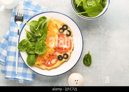 Omelette aux tomates, olives noires, fromage cottage et herbes aux épinards verts sur plaque blanche sur fond de béton gris. Délicieux petit déjeuner. En bonne santé Banque D'Images