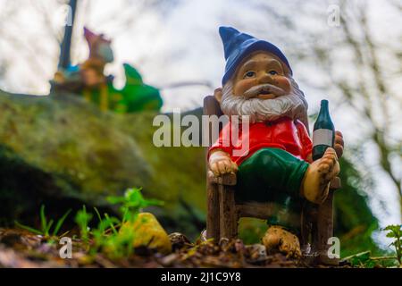 Un gnome de cour se détend dans son fauteuil à bascule avec un gnome de jardin en toile de fond, en faisant monter un lézard. Banque D'Images