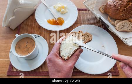 Petit-déjeuner italien avec pain, beurre et confiture. Les mains de l'homme au premier plan répartissent le beurre sur une tranche de pain avec un couteau Banque D'Images