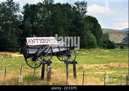 Avec ses intempéries et ses pourriture, ce chariot en bois se trouve au-dessus d'une clôture en métal. Wagon a un panneau fissuré et peeling attaché avec le mot antiquités sur lui. Banque D'Images