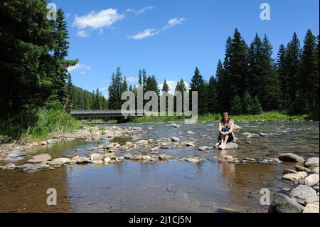 Une jeune femme attrayante est assise sur un rocher au milieu de la rivière Gallatin, au Montana. Elle est souriante et pieds nus avec des jeans roulés. Banque D'Images