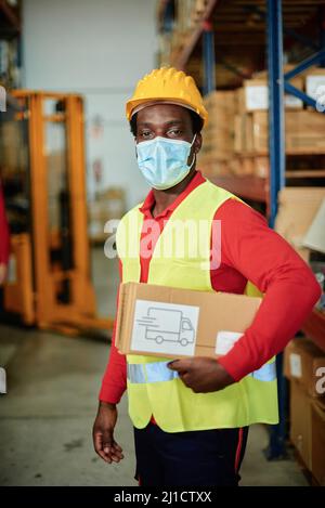 Un travailleur afro-masculin charge une boîte dans un entrepôt de transport portant un masque de sécurité, un gilet réfléchissant et un casque. Concentrez-vous sur les visages Banque D'Images