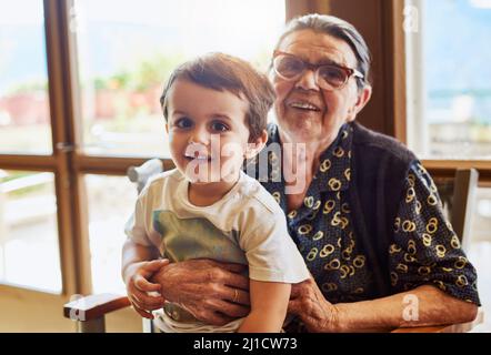 La plus ancienne et la plus jeune de la famille. Portrait d'un petit garçon joyeux assis sur ses grands-mères tout en regardant dans l'appareil photo à la maison. Banque D'Images