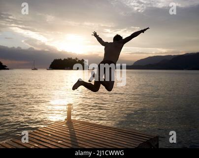 Spontanéité au coucher du soleil. Vue arrière photo d'un jeune homme qui plonge au large de la jetée dans un lac au coucher du soleil. Banque D'Images