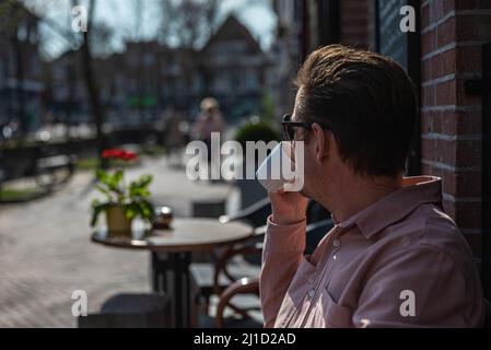 Blanc inreconnaissable homme caucasien dans des vêtements décontractés tient une tasse de café et le boit sur la terrasse en saison de printemps. Banque D'Images
