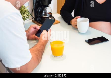 un couple hétérosexuel vénézuélien non reconnaissable autour de la table à la maison buvant du jus et en utilisant le téléphone, femme enceinte avec son mari, Banque D'Images