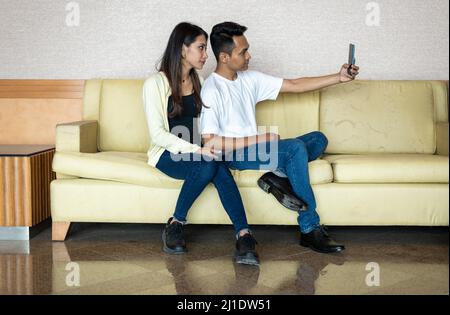 Un couple musulman malais asiatique se réjouit de prendre une photo de selfie à l'aide d'un téléphone à main, tout en étant assis sur un canapé-lit marron clair à la maison Banque D'Images