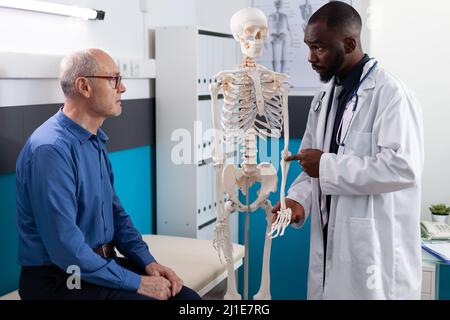 Kinésithérapeute médecin montrant le squelette humain anatomique expliquant la douleur dorsale au patient âgé discutant des symptômes de maladie pendant la physiothérapie médicale dans le bureau de l'hôpital. Traitement de l'ostéoporose Banque D'Images