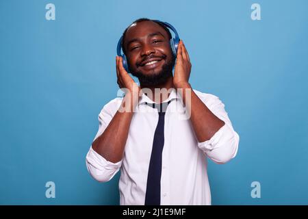 Homme souriant touchant un casque sans fil et se détend en écoutant de la musique à succès sur fond bleu. Homme d'affaires relaxant en chemise blanche, balado audio en streaming dans un casque circum-aural. Banque D'Images