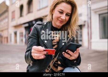 Une femme heureuse s'assoit sur un banc et paie les factures de son mobile intelligent. Banque D'Images
