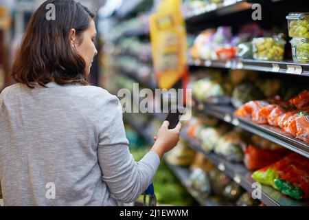 La façon intelligente de faire des achats. Photo rognée d'une femme qui vérifie sa liste d'achats numériques sur son téléphone portable dans une épicerie. Banque D'Images