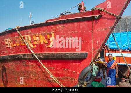 Sous un ciel bleu ensoleillé, une femelle portier se trouve à côté d'un chalutier de pêche rouge ancré au Vieux Port de Mangalore (Mangaluru), Karnataka, Inde du Sud Banque D'Images