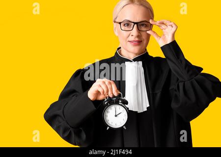 Femme mature juge en robe avec réveil sur fond jaune Banque D'Images