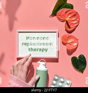 Thérapie de remplacement d'hormone de texte, cadre rose dans la main. Ménopause, concept d'hormonothérapie. Fond rose avec des feuilles exotiques, des fleurs, des pilules Banque D'Images