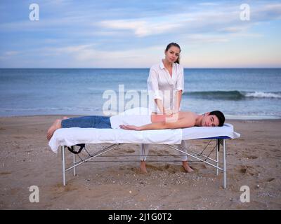 Un jeune thérapeute effectuant un traitement de chiromassage sur un jeune homme pour traiter une blessure lombaire à l'extérieur, tous deux regardant la caméra sur une plage de Valence. Banque D'Images