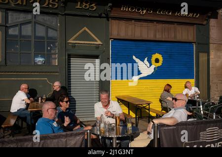 Glasgow, Royaume-Uni, 25th mars 2022. Les gens prennent un verre au soleil devant une fresque murale sur le mur du salon de thé BiddyÕs représentant le drapeau ukrainien et une colombe de paix, peinte pour montrer leur soutien à l'Ukraine dans leur guerre en cours contre l'invasion par la Russie, à l'extrémité est de Glasgow, en Écosse, 25 mars 2022. Crédit photo : Jeremy Sutton-Hibbert/Alay Live News. Banque D'Images