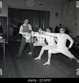 1960s, étudiant historique, masculin portant des culottes, des vestes et des pilmolos traditionnels dans une classe d'escrime, avec instructeur, Université d'Oxford, Angleterre, Royaume-Uni. Ils travaillent sur la poussée des tireurs, un mouvement clé dans le sport, qui est une poussée d'attaque faite avec un pied en avant et la jambe arrière droite et le bras d'épée tendu vers l'avant. Banque D'Images