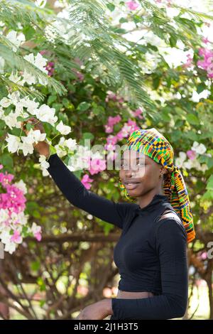 Charmante jeune fille africaine, portant un bandeau coloré, cueillant des fleurs d'un arbre dans un jardin tropical. Banque D'Images