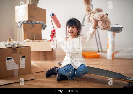 petite fille asiatique souriante, assise sur une planche boostée, tenant un ours en peluche et un rouleau de peinture, avec les mains dans l'air, entourée d'un carton sans emballage bo Banque D'Images