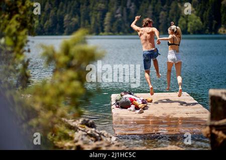Couple tenant les mains sautant sur la jetée en bois au bord du lac. Concept de style de vie, de convivialité, de nature Banque D'Images