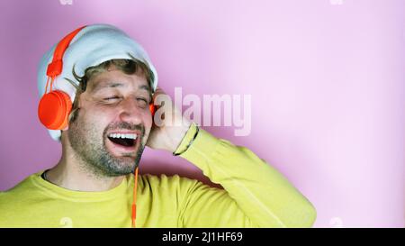 Prise de vue en studio horizontale d'un homme barbu sans souci et gai portant un chapeau blanc, une chemise jaune, pour écouter de la musique avec un volume élevé dans un casque stéréo orange Banque D'Images