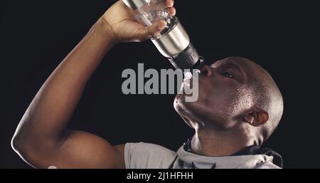 Étancher ma soif avant de faire mes derniers reps. Photo en studio d'un jeune sportif qui boit d'une bouteille d'eau sur un fond sombre. Banque D'Images