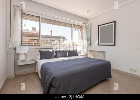 Chambre avec lit double et draps blancs, coussins bleus et couverture bleue assortie sous une grande fenêtre avec vue Banque D'Images