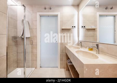 Salle de bains avec lavabo monobloc et deux lavabos en marbre crème sous un miroir rectangulaire avec un cadre fin en bois blanc et une cabine de douche Banque D'Images