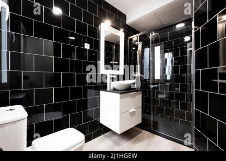 Salle de bains avec murs carrelés de noir, miroir encadré blanc sur le mur et grand meuble en bois Banque D'Images