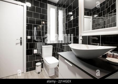 Salle de bains avec murs carrelés de noir, miroir encadré blanc sur le mur et grand meuble lavabo en bois, carrelage en céramique et porte en bois blanc Banque D'Images