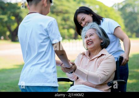 Une grand-mère asiatique heureuse utilise un fauteuil roulant avec sa fille et son petit-enfant dans le parc, le petit-fils est venu rendre visite à une grand-mère âgée et la main. Con Banque D'Images