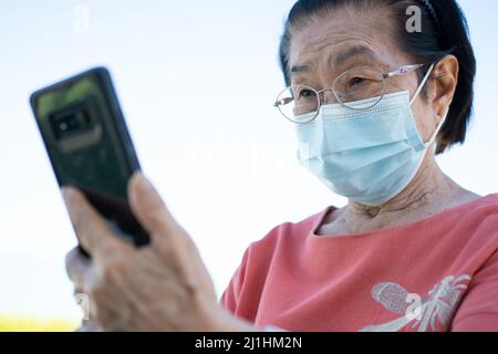 Des personnes âgées asiatiques heureuses portant un masque facial et répondant à un appel vidéo en utilisant son téléphone intelligent dans le parc et en discutant avec la famille, la technologie et les personnes âgées peo Banque D'Images