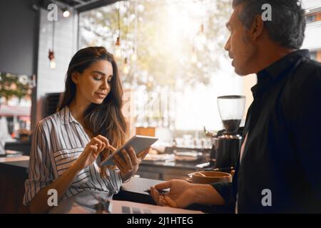 Traitement du paiement. Prise de vue rognée d'une jeune femme attractive traitant un paiement d'un client masculin sur sa tablette tout en travaillant dans son café. Banque D'Images