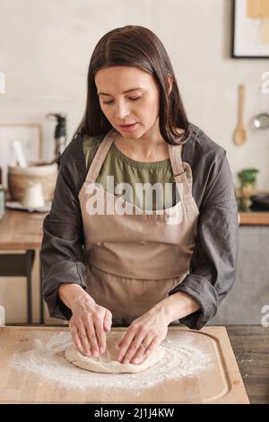 Jeune femme sérieuse en tablier debout à la table avec du bois et pétrir de la pâte tout en cuisinant des petits pains dans la cuisine Banque D'Images