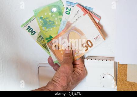 Ancien retraité détenant des billets en main devant un thermostat de chauffage dans un appartement, Allemagne Banque D'Images