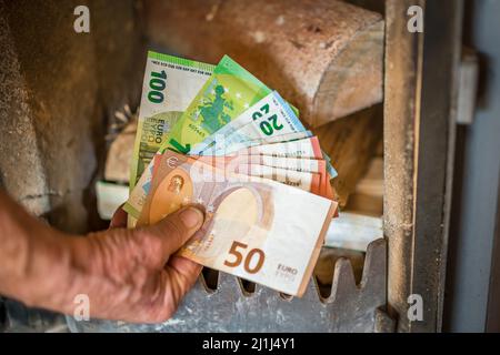 Ancien retraité tenant des billets en main devant un poêle à bois avec du bois dans l'appartement, Allemagne Banque D'Images