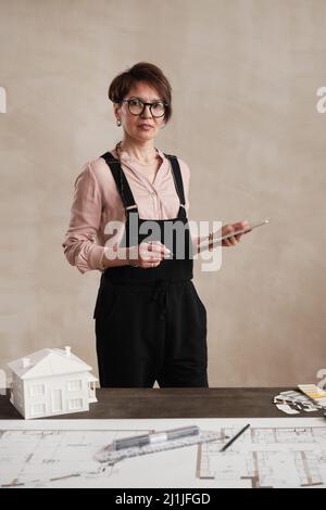 Portrait d'une femme d'architecte caucasienne souriante en lunettes debout avec une tablette au bureau pleine d'outils de conception Banque D'Images