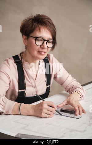 Concepteur architectural caucasien mature positif en lunettes assises à la table et ligne de dessin à l'aide d'une règle Banque D'Images