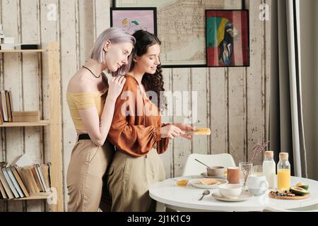 Une jeune femme soigneuse met de la confiture sur des toasts pour petite amie pendant qu'ils prennent le petit déjeuner ensemble à la maison Banque D'Images