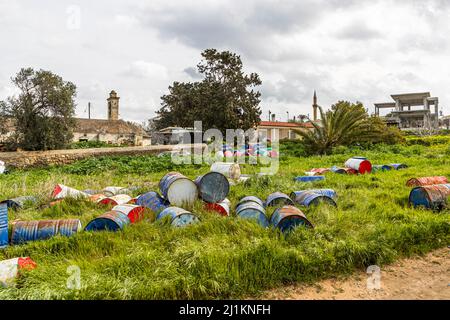 Des fûts d'huile sont dispersés sur un champ en jachère à la périphérie d'Akdeniz, avec des maisons, un minaret et un bâtiment en béton inachevé visibles en arrière-plan. Akdeniz (turc pour «Mer Méditerranée») offre un aperçu de la vie rurale typique de Chypre, République turque de Chypre du Nord (RTNC). Banque D'Images
