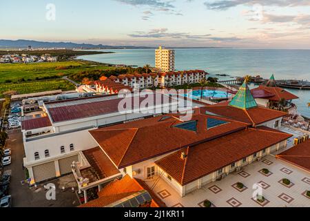 L'hôtel Salamis Bay Conti s'étend vu du dessus, avec des toits de tuiles rouges, une piscine et un pavillon de restaurant à dôme vert sur le rivage près de Yeni Boğaziçi, République turque de Chypre du Nord (RTNC). Une tour d'appartements s'élève en arrière-plan, et la côte continue vers Famagusta. Une ruine d'hôtel voisin se trouve juste à côté sur la plage. Banque D'Images
