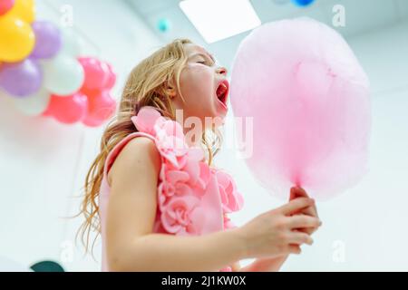 Petite fille avec de longs cheveux justes ondulés tenant des bonbons de coton avec la bouche large ouvert Banque D'Images