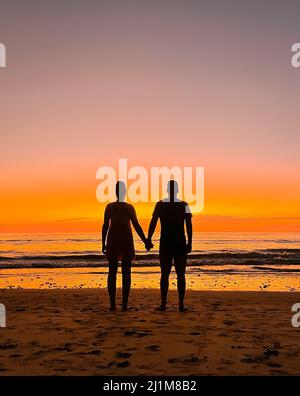 Silhouette de couple tenant les mains en regardant le coucher du soleil sur la plage. Banque D'Images