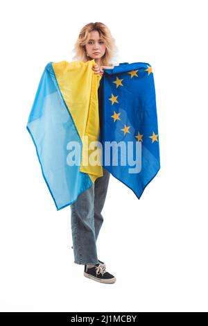 Fière et bouleversée, en 20s, une femme blanche aux cheveux blonds tenant ensemble les drapeaux ukrainiens et européens. L'Europe pour l'Ukraine. Studio vertical tourné sur fond blanc. Photo de haute qualité Banque D'Images