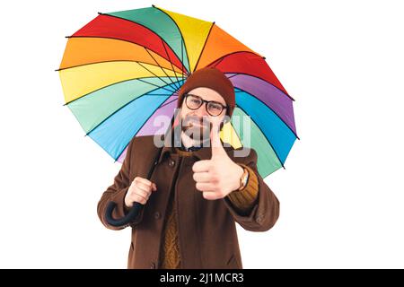 Portrait d'un jeune homme caucasien attrayant tenant un parapluie multicolore et montrant le pouce jusqu'à la caméra positivité optimisme concept marron décontracté tenue isolée blanc arrière-plan studio tourné . Photo de haute qualité Banque D'Images