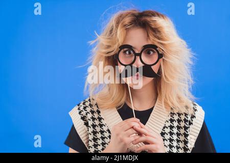 Amusant goofy adolescent caucasien tenant la fausse moustache noire près de son visage, isolé sur fond bleu. Photo de haute qualité Banque D'Images