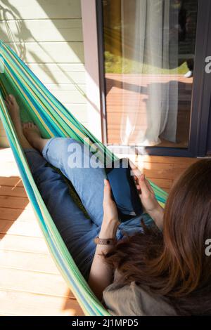 Vue latérale d'une jeune femme balance dans un hamac vert près de la maison de campagne et utilise un smartphone. Banque D'Images