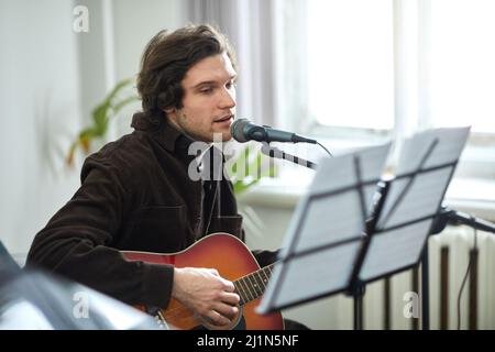 Jeune homme regardant la musique de feuille et chantant une chanson au microphone tout en jouant la guitare en studio Banque D'Images