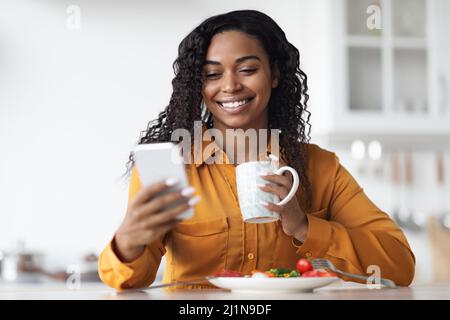 Femme noire souriante qui vérifie son smartphone tout en prenant le petit déjeuner Banque D'Images