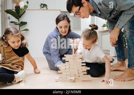 Père délicat soufflant sur le mur de jenga, tandis que son fils tirant une tuile hors de lui. Parents et enfants assis sur le sol. Banque D'Images